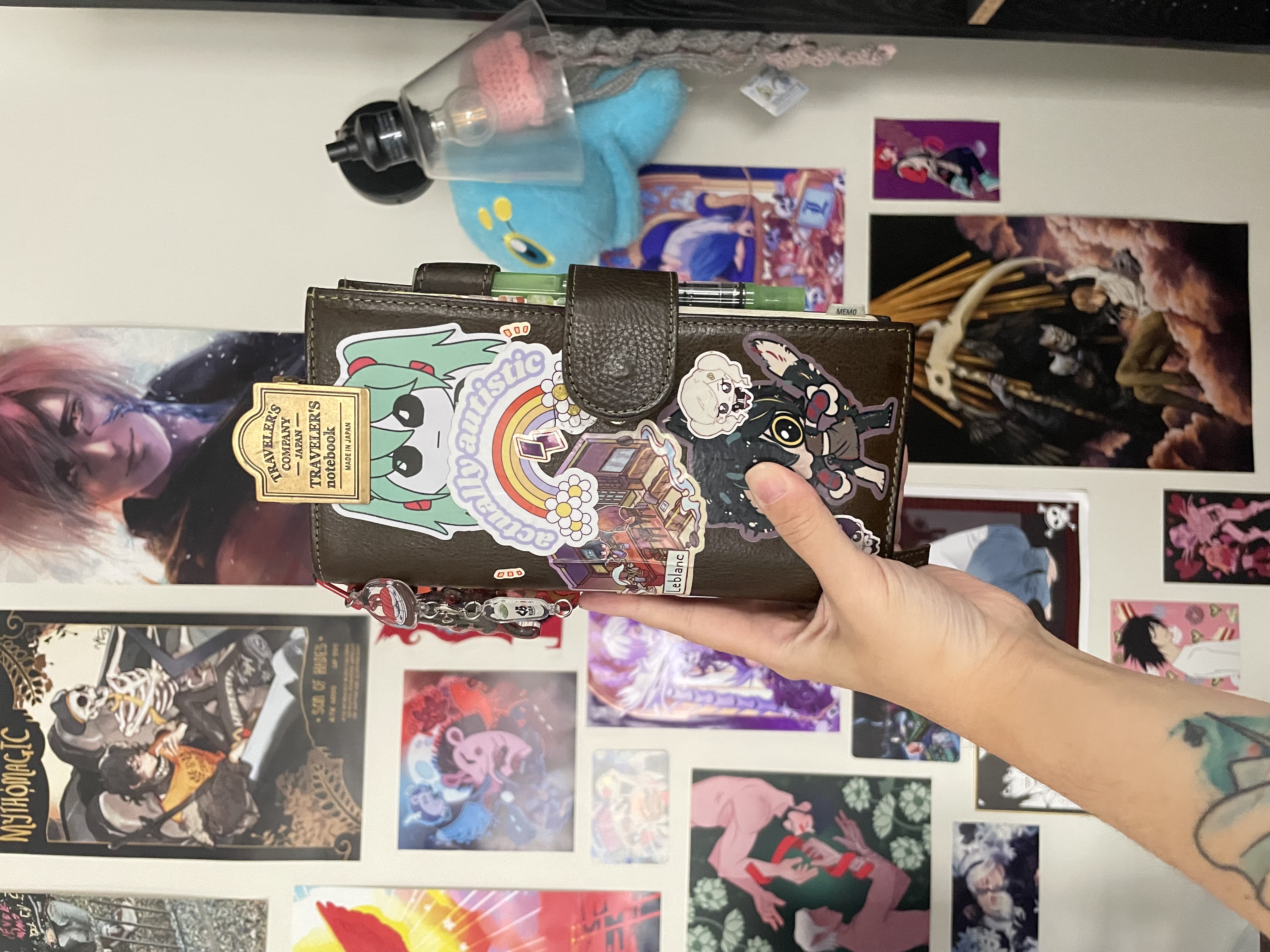 an olive green moterm-brand weeks cover with a snap closure. it has a metal traveler's company clip and a few charms on it, one visibly being a kura sushi charm. it's covered in stickers: a tbh creature drawn as miku, a rainbow with the words 'actually autstic' over it, an akeshu persona 5 sticker of them hanging out in leblanc together, a chibi sticker of ann and yusuke from persona 5, and one of izutsumi from dungeon meshi looking feral. in the cover's pen loop, there is a TWSBI glow-green fountain pen. yes, it glows in the dark.